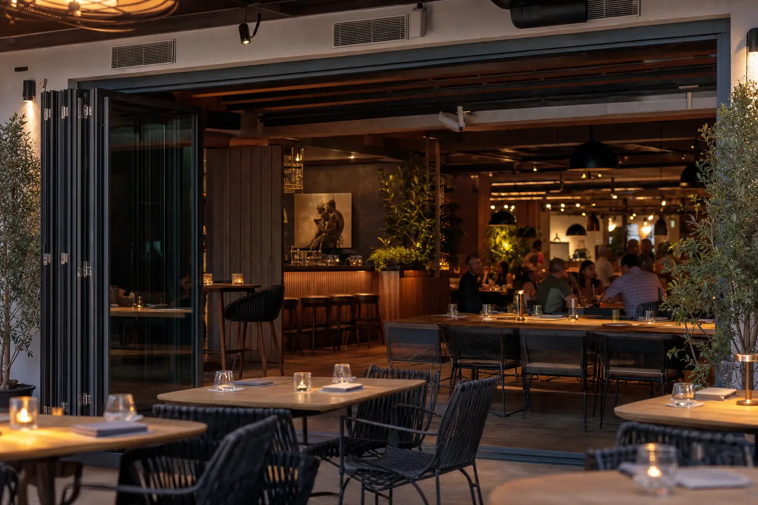Restaurant terrace at dusk with a folding glass wall stacked fully open to the dining room