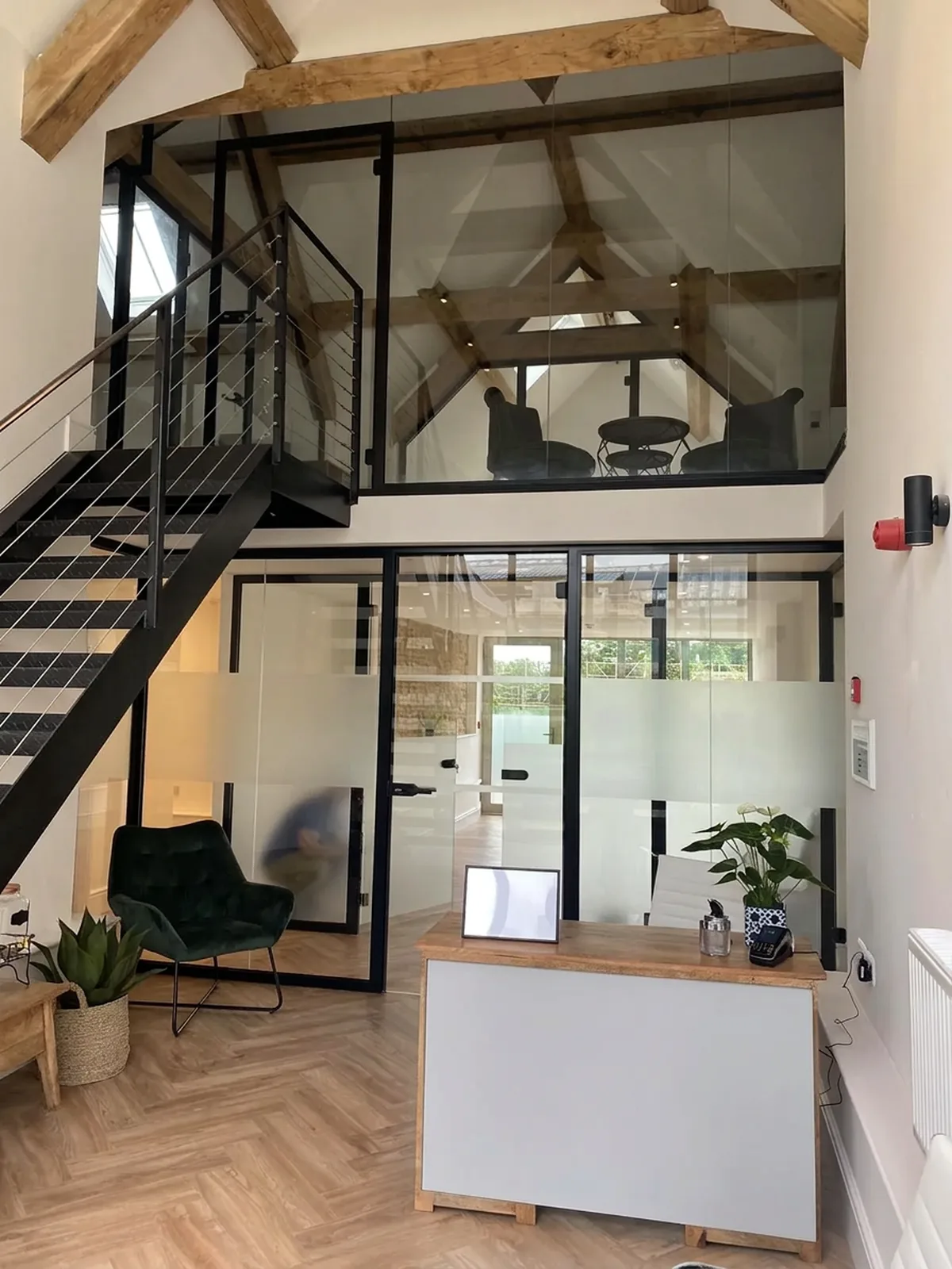 Double-height reception space enclosed by black-framed glazed partitions, with a glazed mezzanine balustrade above