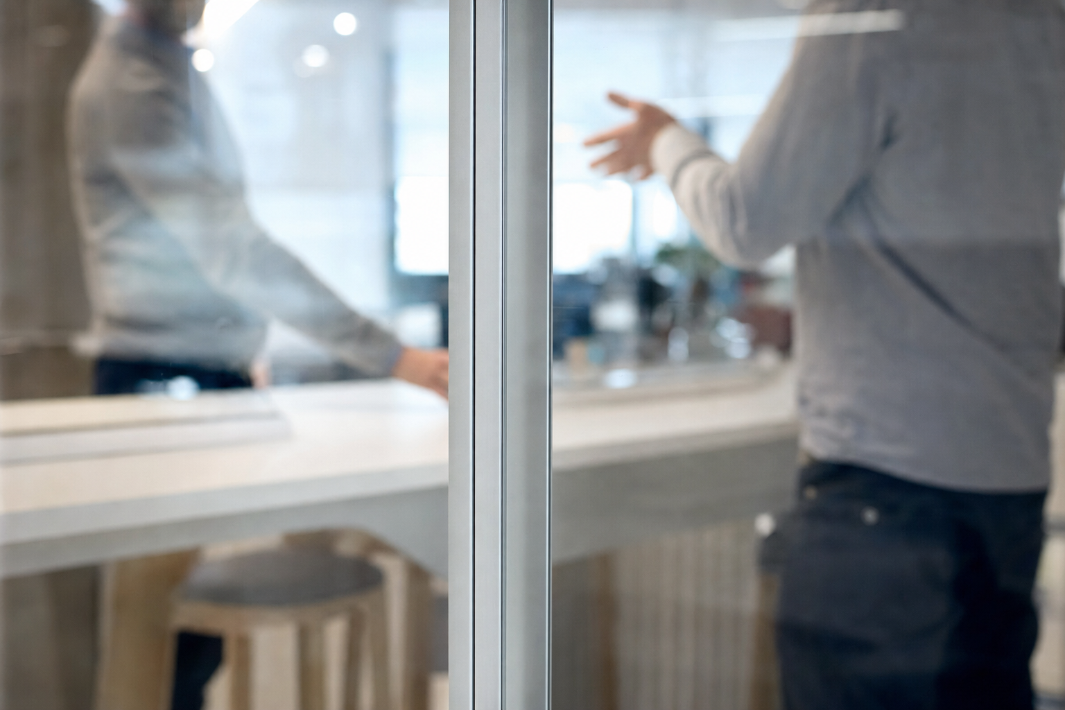 Close view through a slim-framed glass partition, with two people talking at a bench beyond